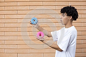 Surprised African-American guy standing sideways, wearing a white T-shirt, holding a pink and blue donut