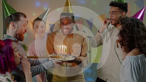 Surprised African American man blowing candles on birthday cake while friends laughing and clapping hands at home party