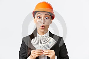 Surprised and amazed asian female engineer in safety helmet and business suit, holding money and looking astounded with
