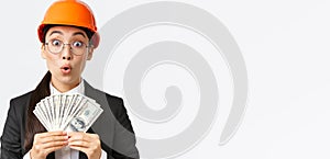 Surprised and amazed asian female engineer in safety helmet and business suit, holding money and looking astounded with