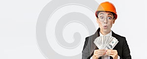 Surprised and amazed asian female engineer in safety helmet and business suit, holding money and looking astounded with