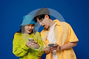 Surprised asian couple looking at mobile phone screen isolated over blue background