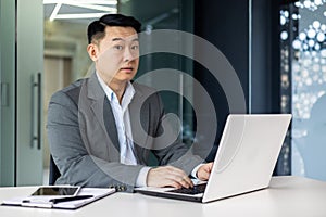 Surprised asian man looking at camera, mature adult boss businessman working inside office with laptop while sitting at