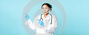 Surprised asian woman doctor or nurse, pointing fingers left and looking amazed at advertisement, copy space, standing