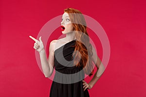 Surprised and astounded young redhead woman in elegant party black dress, drop jaw gaspins as staring and pointing