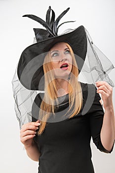 Surprised attractive blond woman in black witch hat with green eyes looks up. Young witch, vertical frame. Halloween party, white