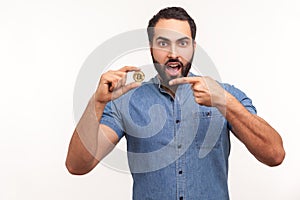 Surprised bearded man pointing finger at golden bitcoin cryptocurrency, looking at camera with astonishment, shocked with price