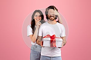 Surprised bearded man in a white shirt unknowingly holds a gift with a red ribbon as a smiling woman