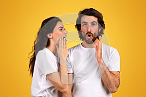 Surprised bearded man with wide eyes and an open mouth holds his hands up while a woman whispers into his ear