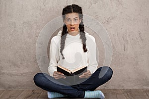 Surprised beautiful african girl holding book, sitting over beige background.