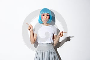 Surprised beautiful asian girl in blue short wig, standing over white background and pointing fingers sideways with