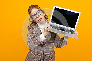Surprised beautiful girl in a classic jacket shows a laptop with a mock-up display forward on a yellow background with