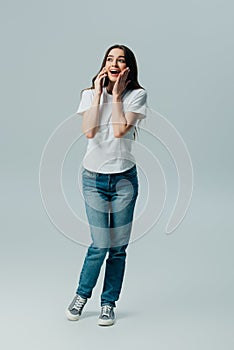 Surprised beautiful girl in white t-shirt and jeans talking on smartphone isolated on grey
