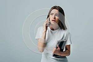 Surprised beautiful girl in white t-shirt talking on smartphone and holding notebook isolated on grey