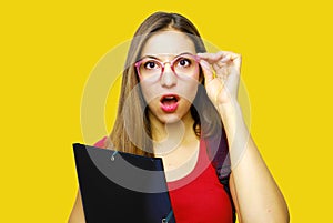 Surprised beautiful student girl. Young shocked student woman holding folder on yellow background