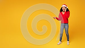 Surprised black lady in Santa hat points at space, yellow background