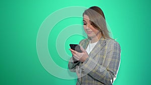 Surprised blonde girl texting on mobile phone while standing isolated over green background