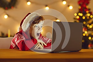 Surprised boy in Christmas festive clothing and hat watching movie online on laptop