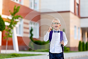 Surprised boy schoolboy blond in glasses with a backpack stands at the school and shows a thumbs up. Day of knowledge
