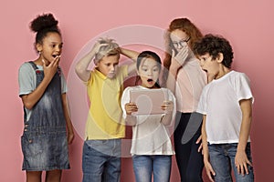 Surprised boys and girls looking at tablet computer together, learning shocking news over pink background