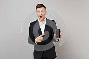 Surprised business man keeping mouth open, pointing index finger on mobile phone with blank empty screen isolated on