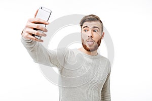 Surprised casual young man taking selfie and showing tongue over white background