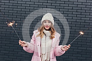 Surprised caucasian girl in pink coat posing with bengal lights. Outdoor photo of blithesome blonde lady holding