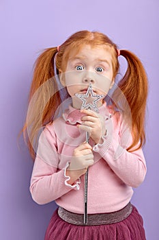 Surprised caucasian redhead girl holding magic wand, looking at camera, pastel purple studio wall, copy space