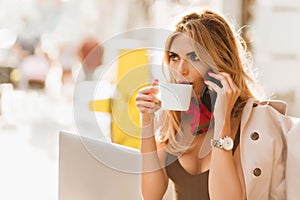 Surprised charming girl drinking hot beverage and listening friend on phone. Attractive business-woman enjoying coffee