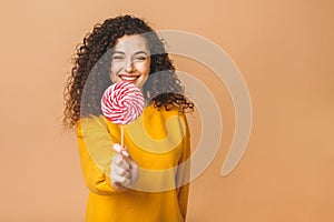 Surprised curly girl eating lollipop. Beauty Model woman holding pink sweet colorful lollipop candy, isolated on beige background