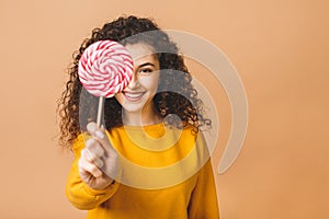 Surprised curly girl eating lollipop. Beauty Model woman holding pink sweet colorful lollipop candy, isolated on beige background