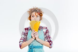 Surprised cute housewife in kitchen apron hiding behind uncooked spagetti