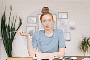 Surprised disappointed redhead young woman student is sitting at a table with books and a workbook.