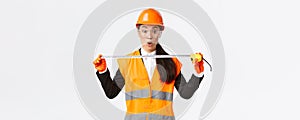 Surprised and excited asian female construction engineer, industrial worker in safety helmet and uniform, measuring