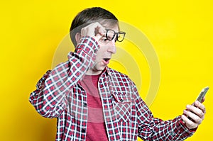 Surprised and excited man with open mouth in red checkered shirt taking off glasses on forehead and looking at smartphone on yello
