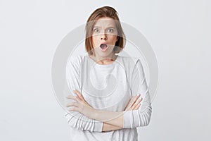 Surprised excited young woman in longsleeve with opened mouth standing with arms crossed and shouting over white background