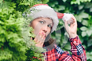 Surprised face woman in a santa claus red hat looks away through green fir tree branches in the forest. Christmas coming, sale