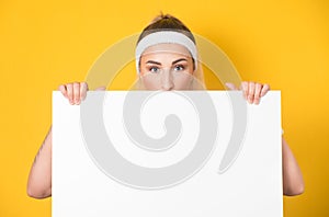 Surprised face of young women behind white board,  on yellow background. One fitness girl showing blank empty paper