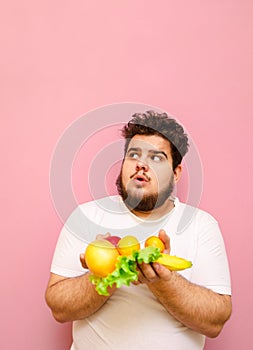 Surprised fat man on diet standing with fruits in hands on pink background and looking up at copy space with shocked face.