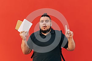 Surprised fat young male student with beard stands on red background with book and notebook in hand and shows finger upward on