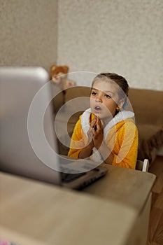 Surprised girl of 7 in a yellow dressing gown sits in front of her laptop at home