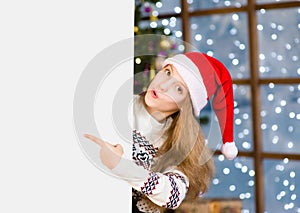 Surprised girl in christmas hat peeking and pointing at empty board