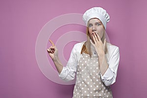 Surprised girl cook in kitchen clothes closes her mouth with her hands and points her finger at a place for text on a pink