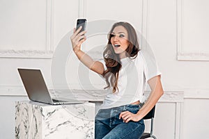 Surprised girl freelancer with a laptop and phone in her hand makes selfie. Young woman with a white T-shirt and jeans