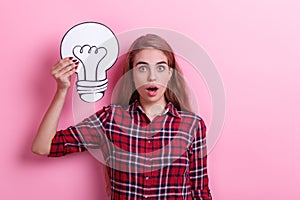 Surprised girl, holding a white paper light bulb. Concept of the idea. On a pink background.
