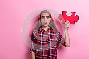 A surprised girl, holds a large puzzle on raised hand with mouth open. On a pink background.