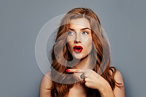 Surprised girl pointing. Excited redhead woman pointing on blue background with copy space