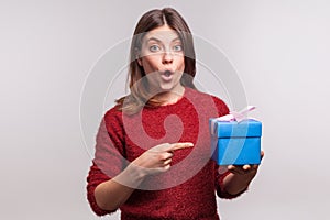 Surprised girl in shaggy sweater pointing decorated wrapped gift box and looking at camera with amazed expression photo