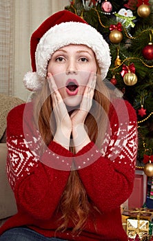 Surprised girl touch face. Home interior with christmas decoration, fir tree and gifts. New year eve and winter holiday concept.