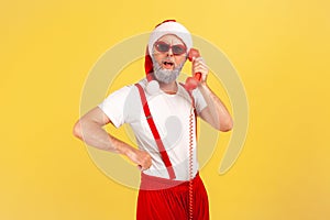 Surprised grey bearded man in santa claus costume and trendy sunglasses calling holding landline telephone handset, congratulating